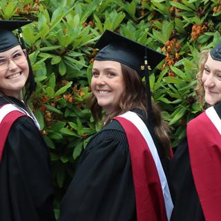 Three students wearing caps and gowns in front of a bush