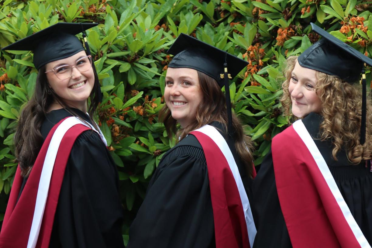 Three students wearing caps and gowns in front of a bush