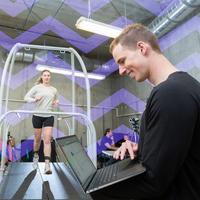 A young man working in a kinesiology lab with a person running on a treadmill.